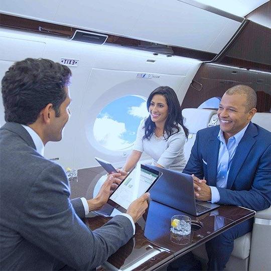 Two men and one woman sitting at a table in a private jet, using tablets and a laptop connected with WiFi for private aircraft