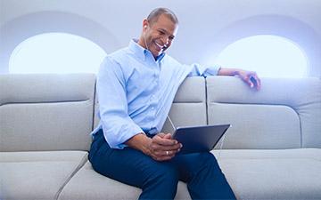 Smiling man sitting in a private jet wtih private jet internet, dressed in a blue button up shirt looking at a tablet